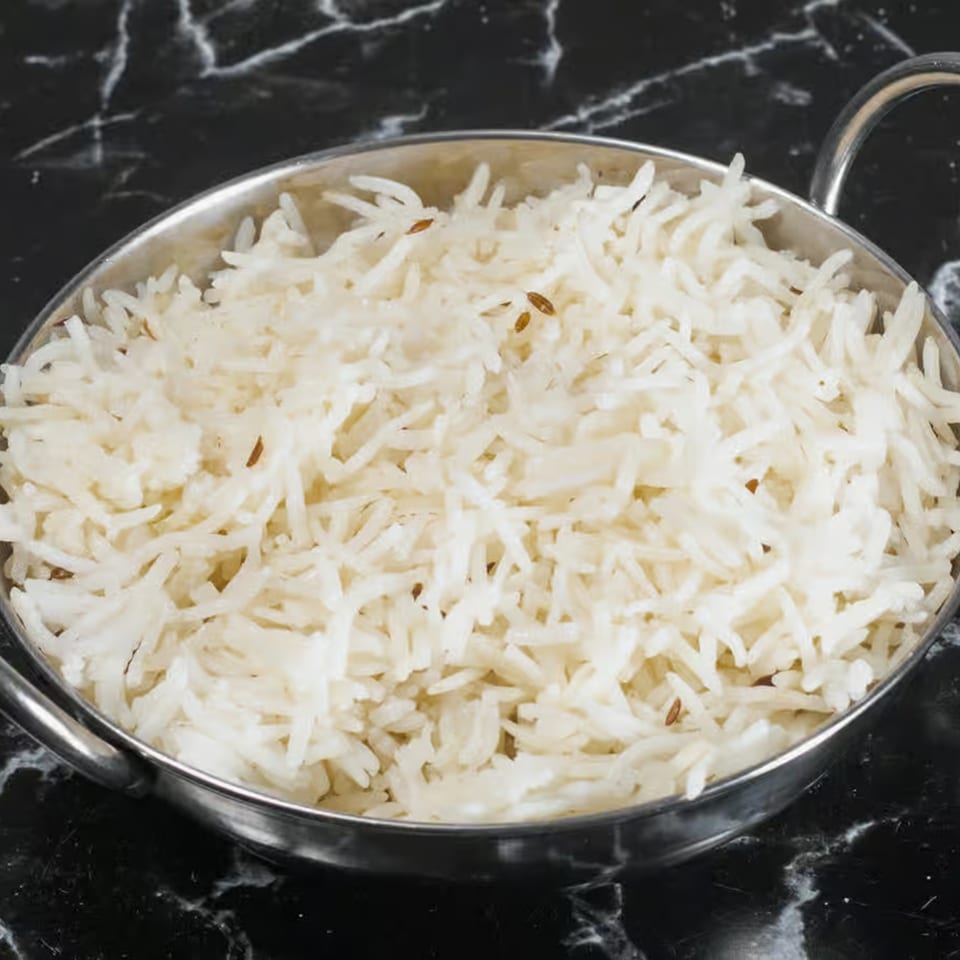 A silver bowl filled with fluffy white basmati rice, sprinkled with cumin seeds, on a dark marble surface.
